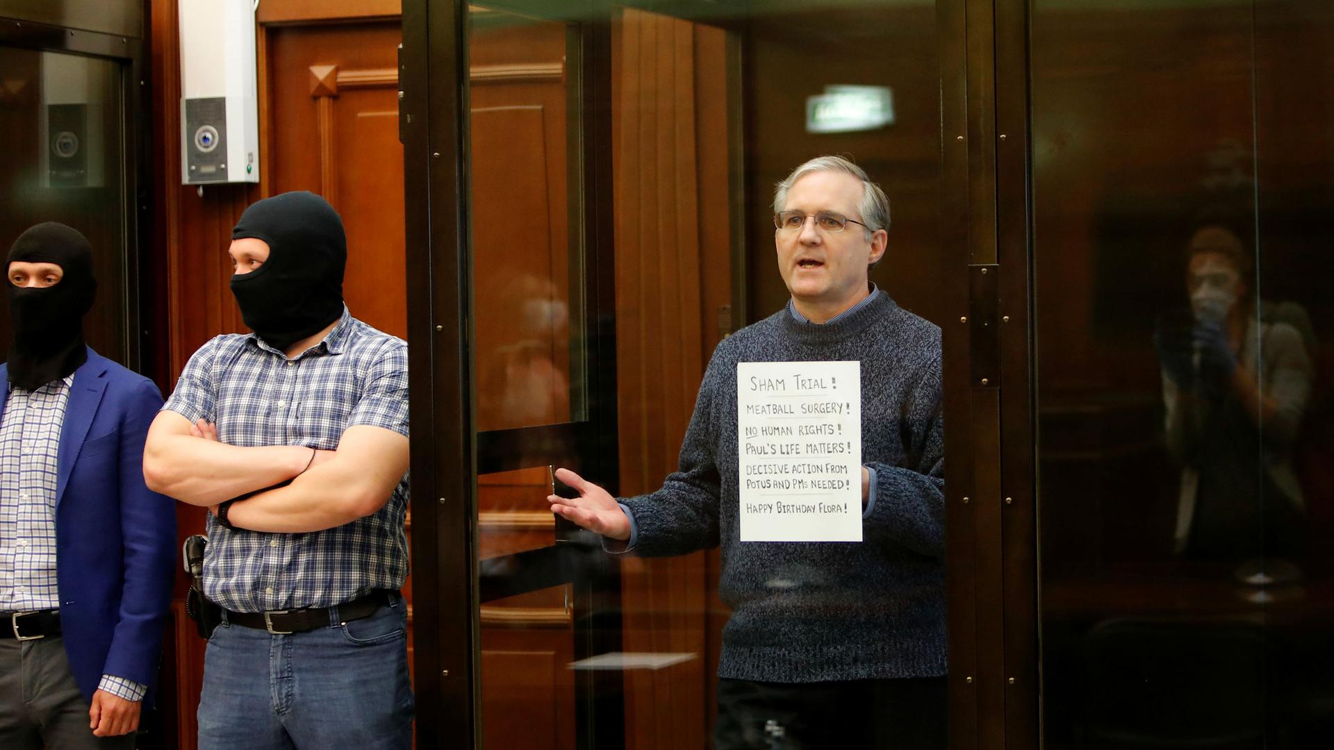 Paul Whelan is shown standing in a glass enclosed box wearing a sweater with two men standing out side of the glass wearing balaclavas.