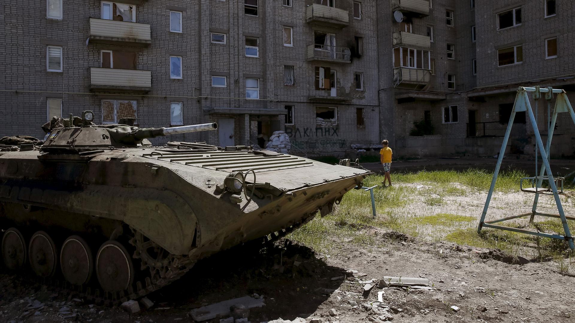 A boy walks near a Soviet-era apartment building, with a tank in the foreground