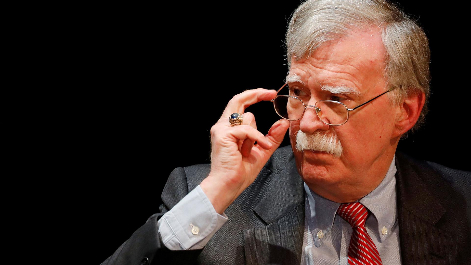 Former US national security advisor John Bolton is shown wearing a stripped suit and red tie and adjusting his glasses.