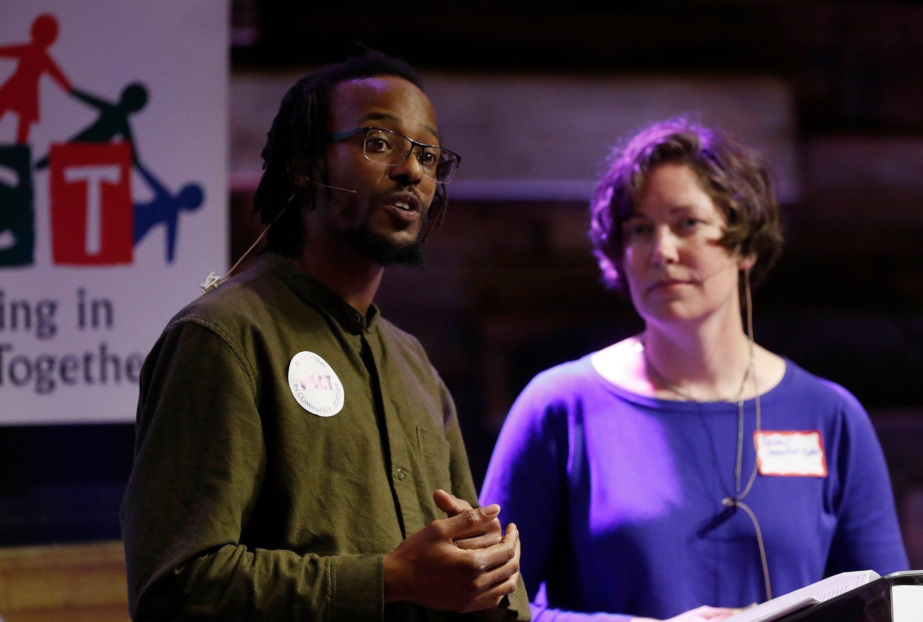 A Black, male community organizer wearing a green shirt speaks next to a white woman wearing a purple shirt at a meeting of People Acting in Community Together in San Jose, California.