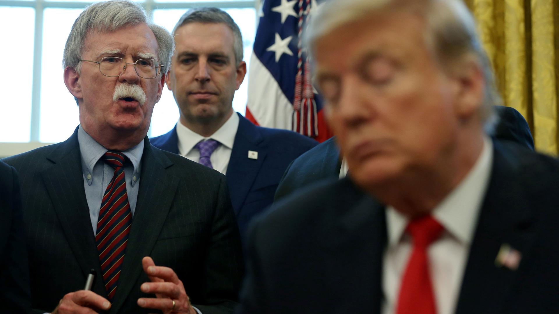 John Bolton is shown speaking in distance with US President Donald Trump in the nearground in soft focus.