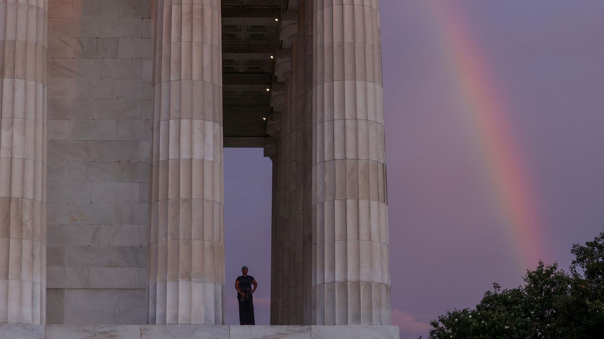 A side view of the Lincoln Memorial shows a woman standing near the large pillars and a rainbow in the distance.