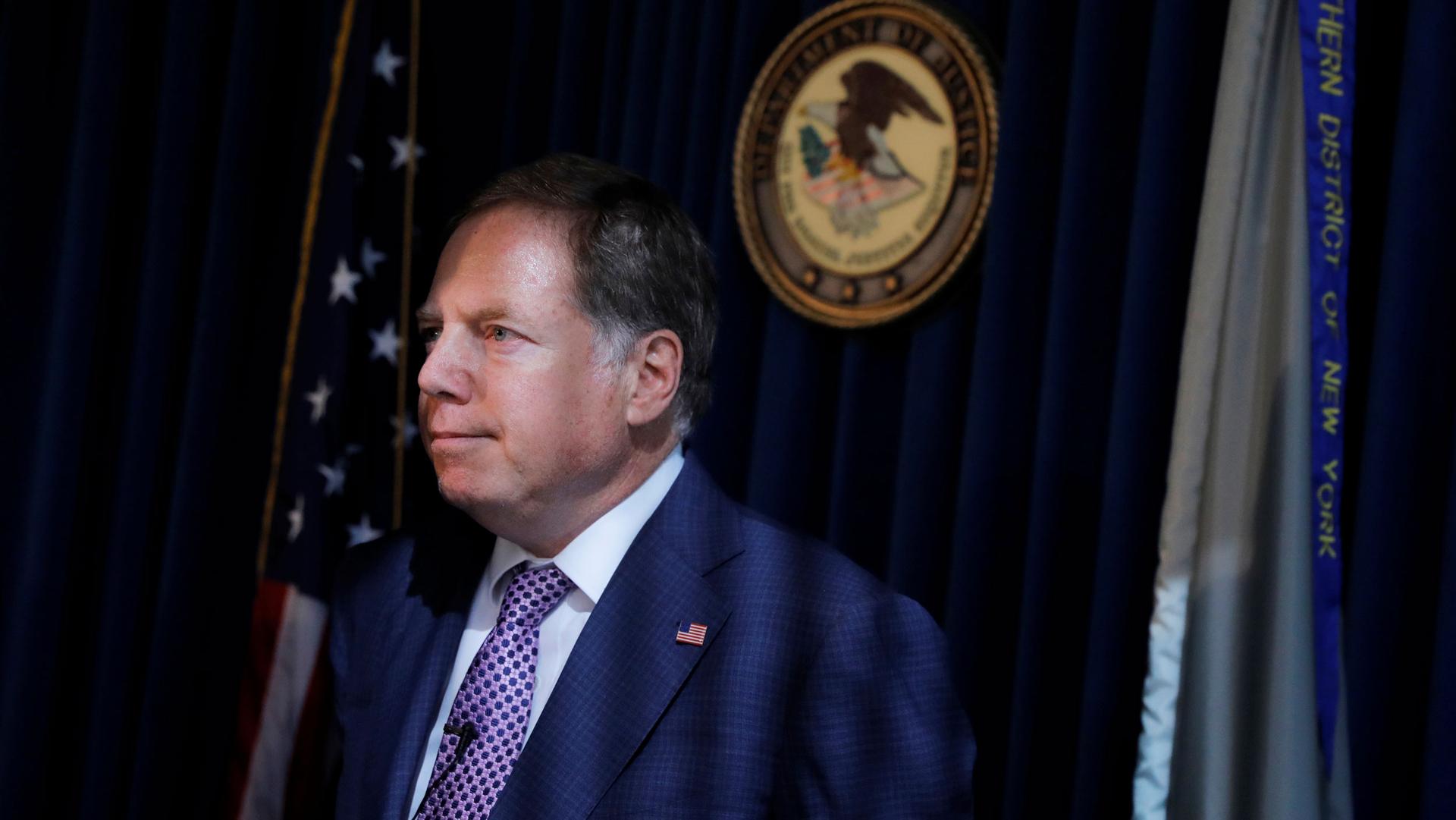 US Attorney for the Southern District Geoffrey Berman is shown wearing a dark suit and purple tie with a US flag behind him.