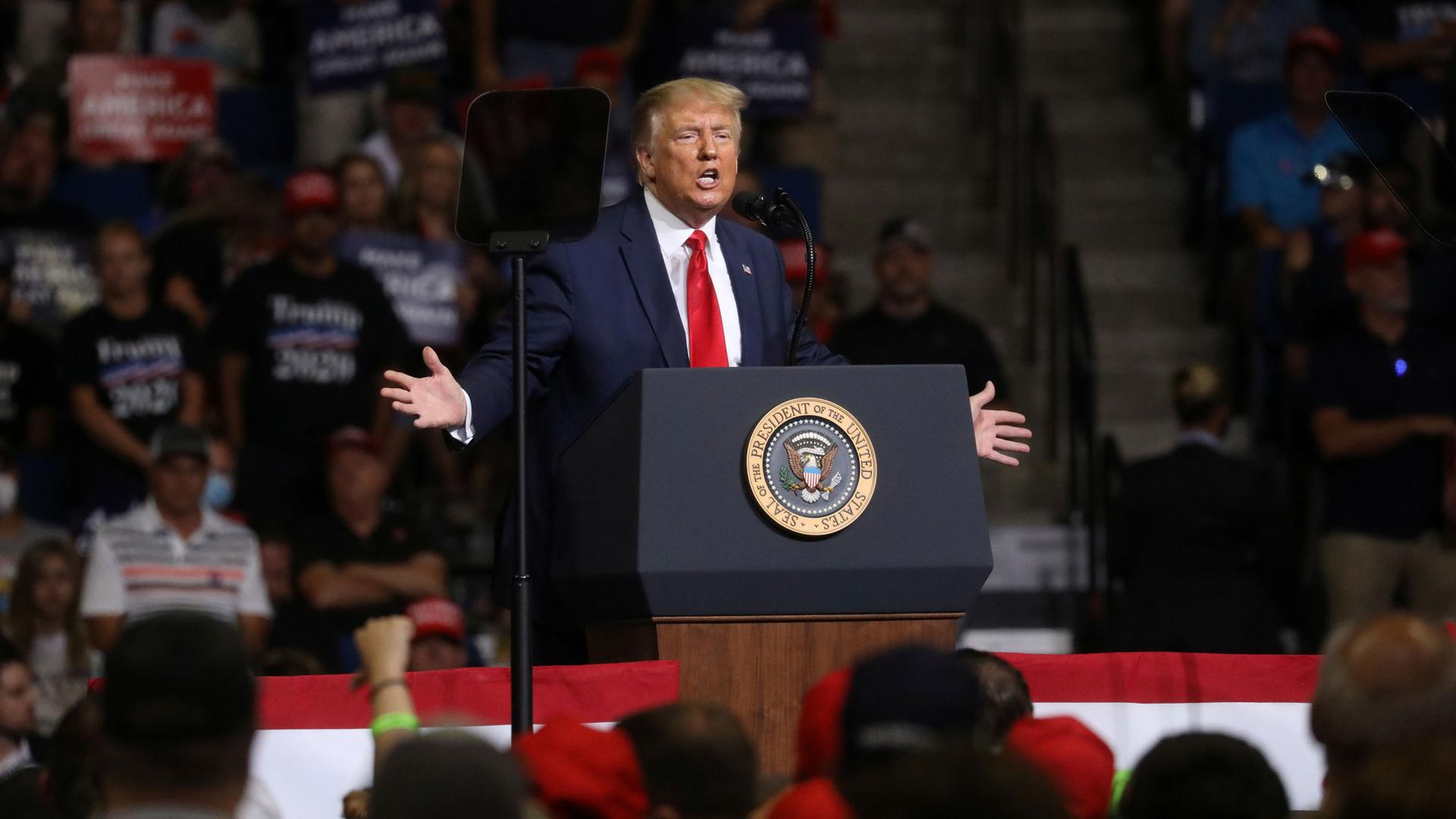 US President Donald Trump is shown standing with his arms outstretched, behind a podium.