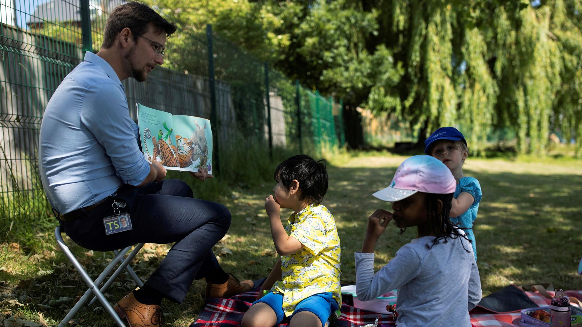 Three children sit on the ground as a male teacher reads a picture book