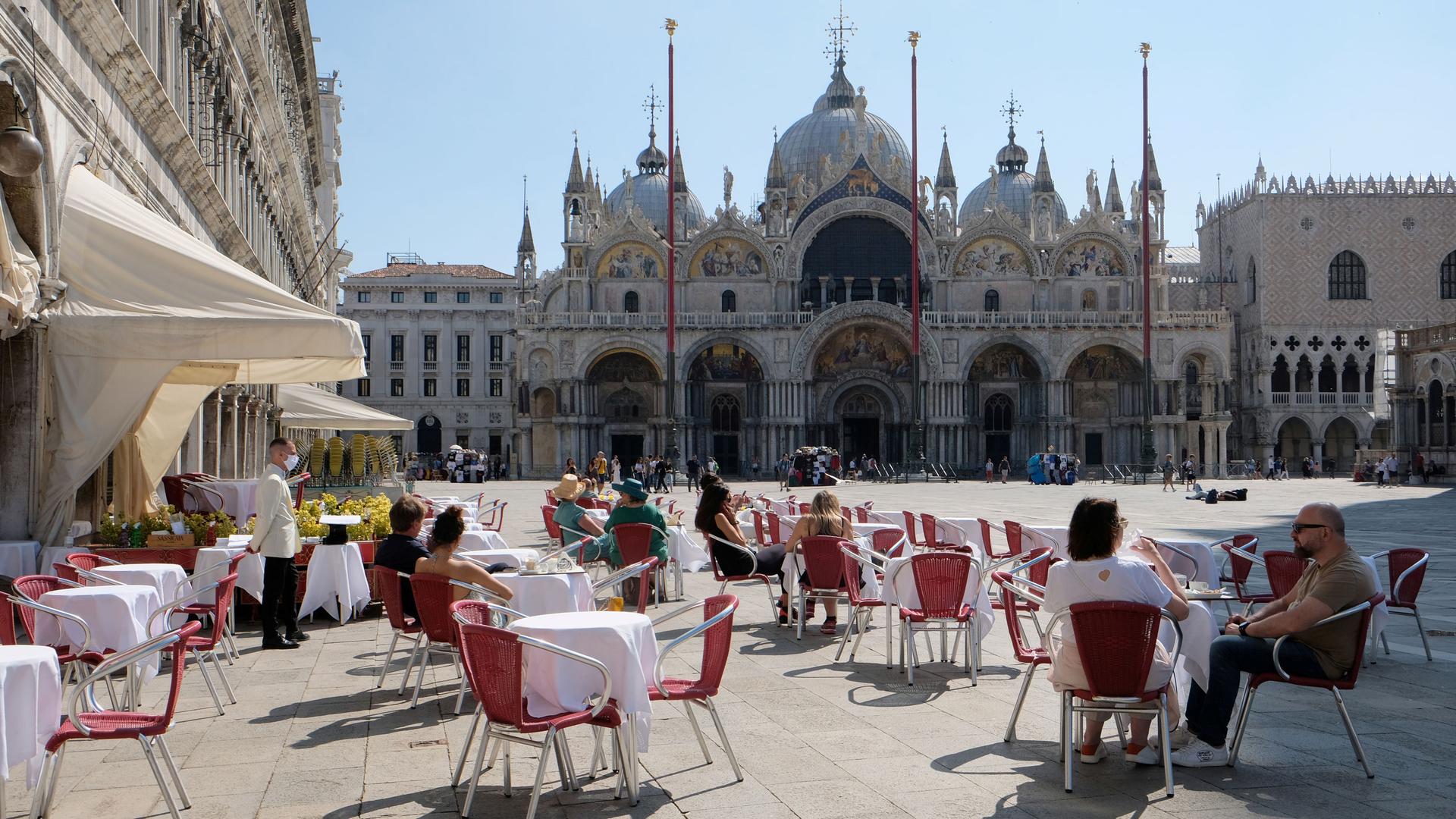 Several cafe tables are shown in St. Mark's Square with the St. Mark's Basilica in the distance.