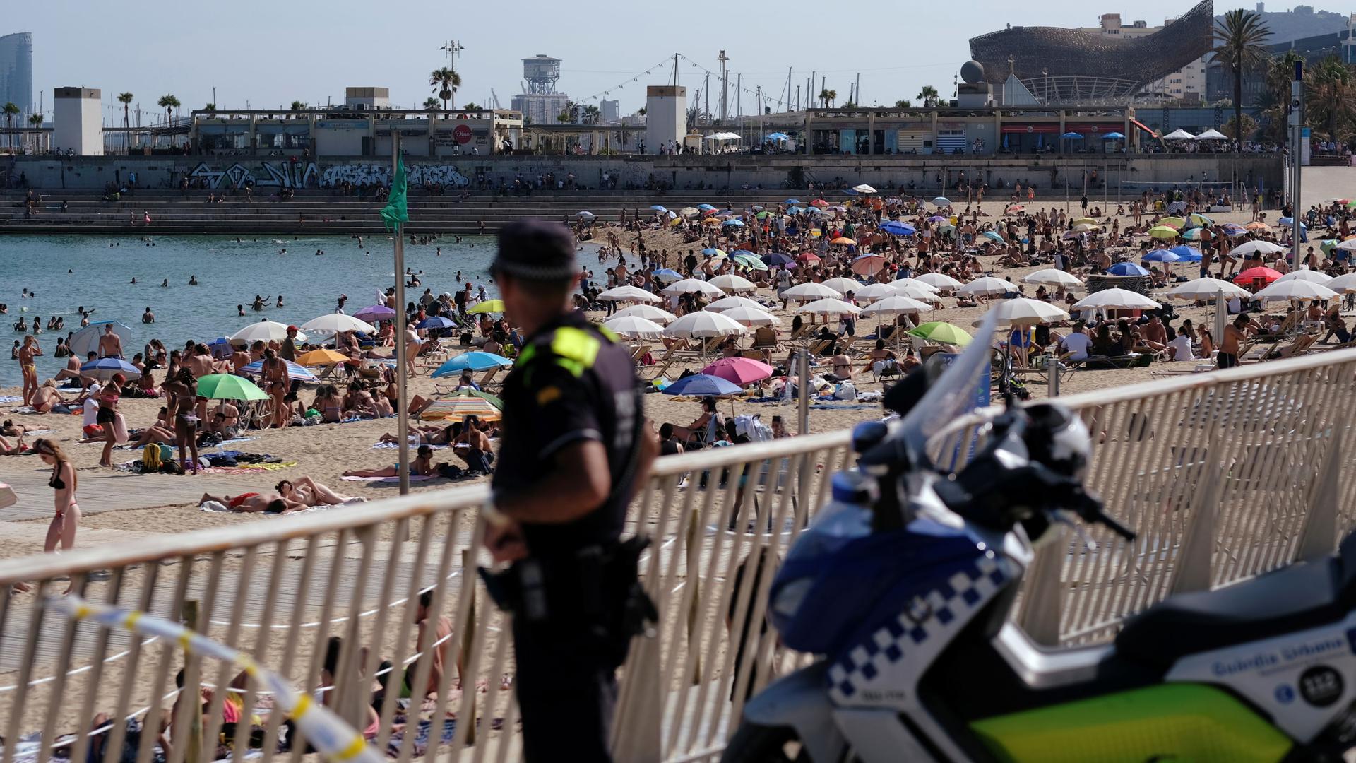 A police officer is shown in the nearground in soft focus with hundreds of beachgoers line the coast with umbrellas.