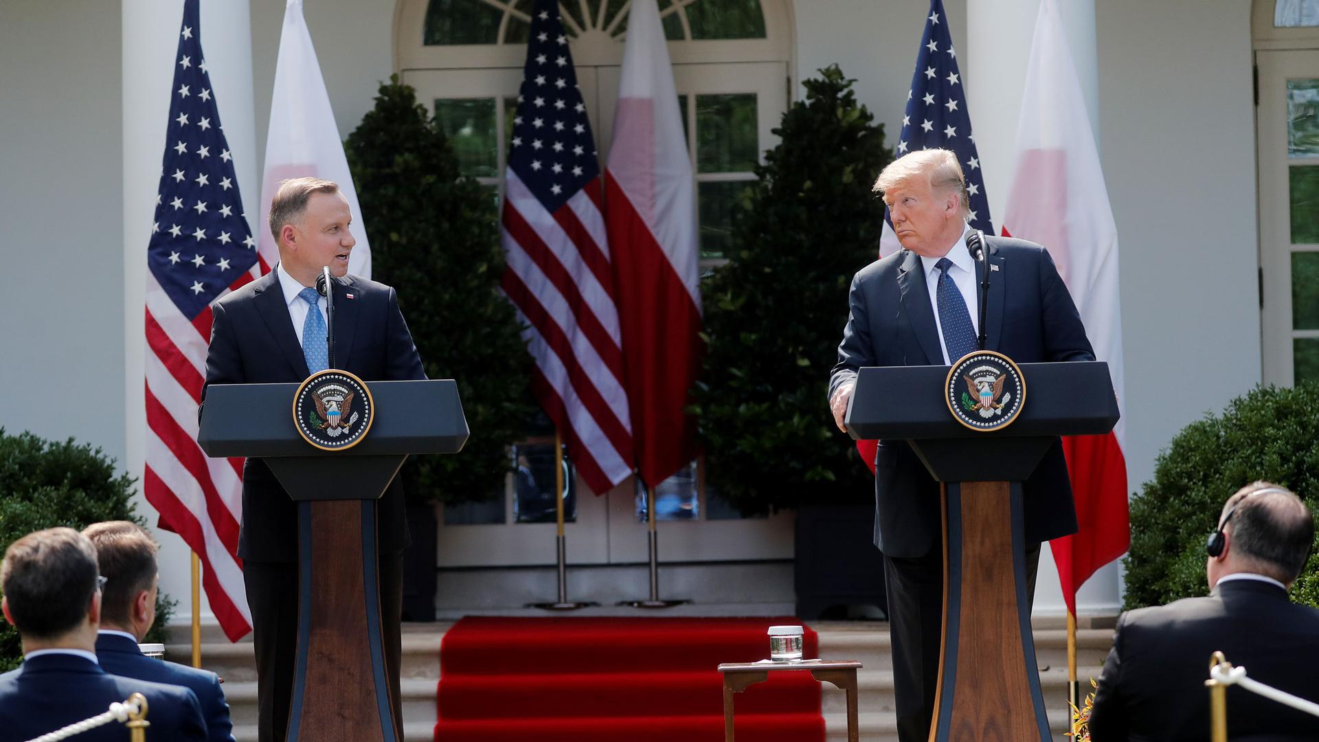 Two white men in suits stand at podiums in front of the US and Polish flags