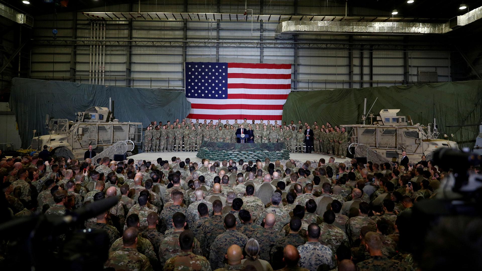 A crowd of US service members stand in front of Trump, with a large US flag in the background