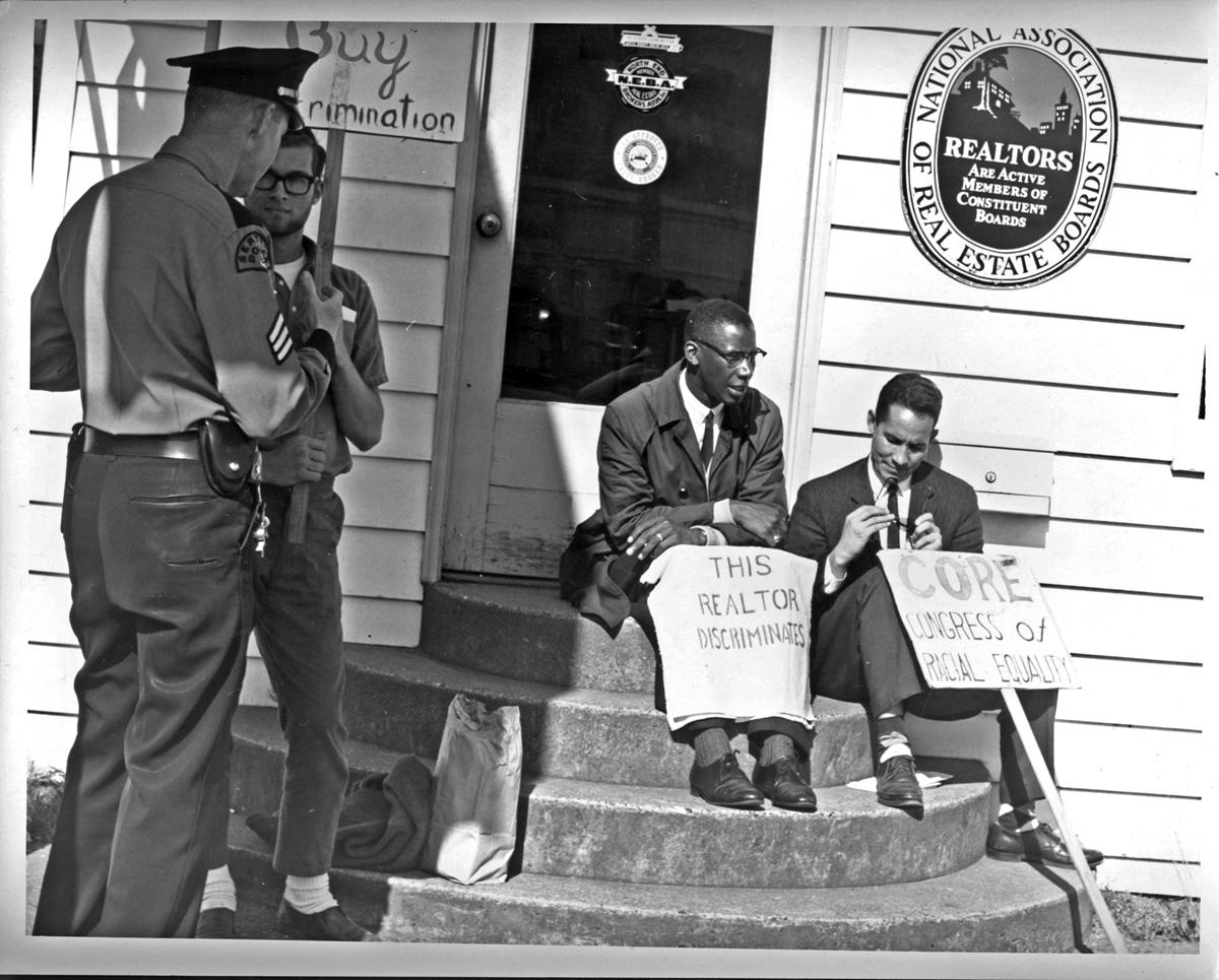 Two black men sit on steps with two white police offers standing by them.