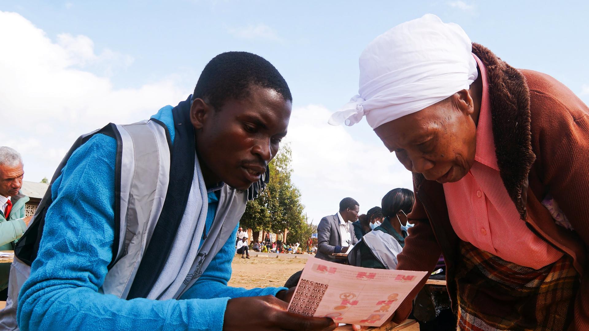 A man in Malawi wearing a blue shirt explains a document to an elderly woman wearing a white head wrap outside 