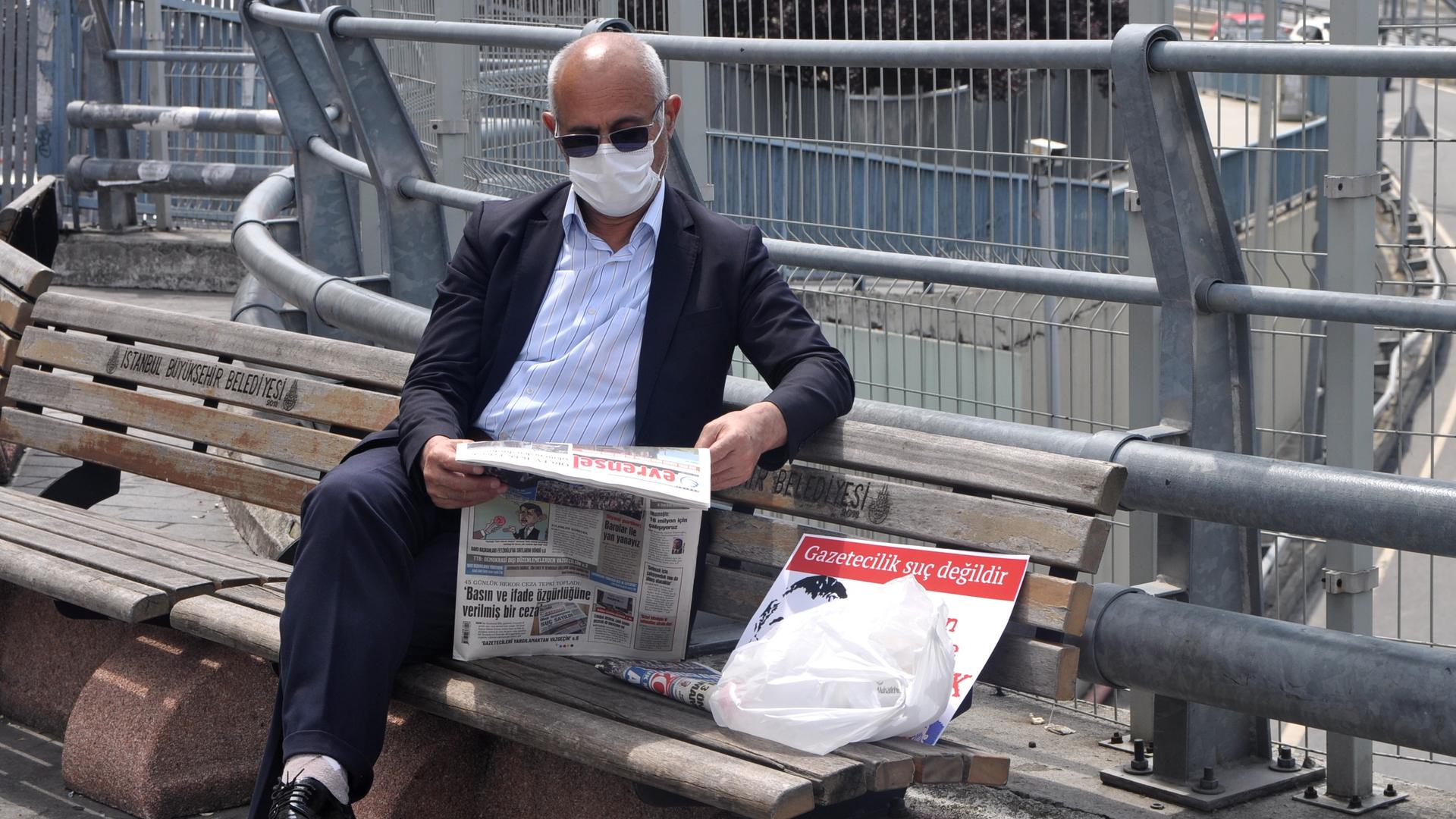 A man in a suit reads a newspaper on a bench outside 