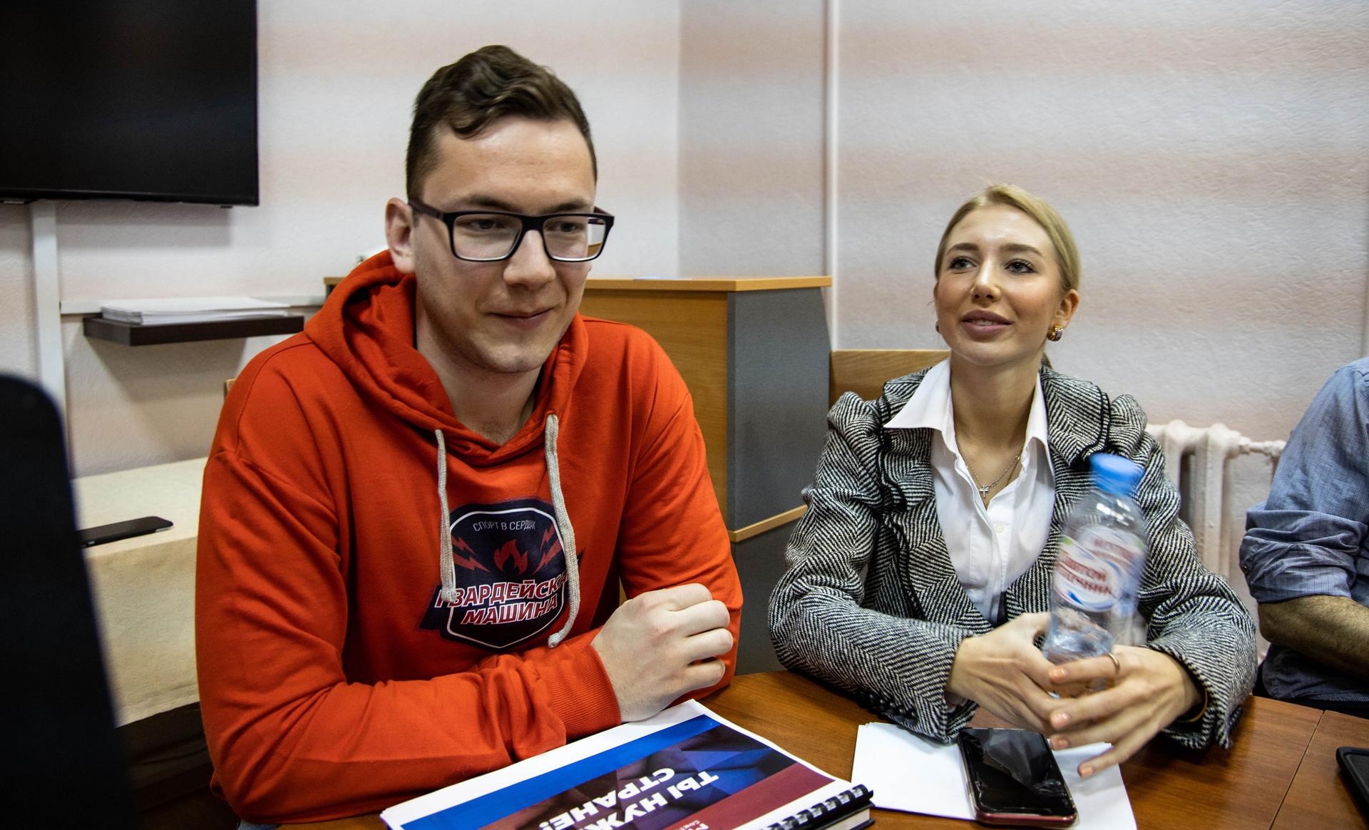 Two young people sit at a table. One young man is wearing an orange hoodie with Russian words on it.