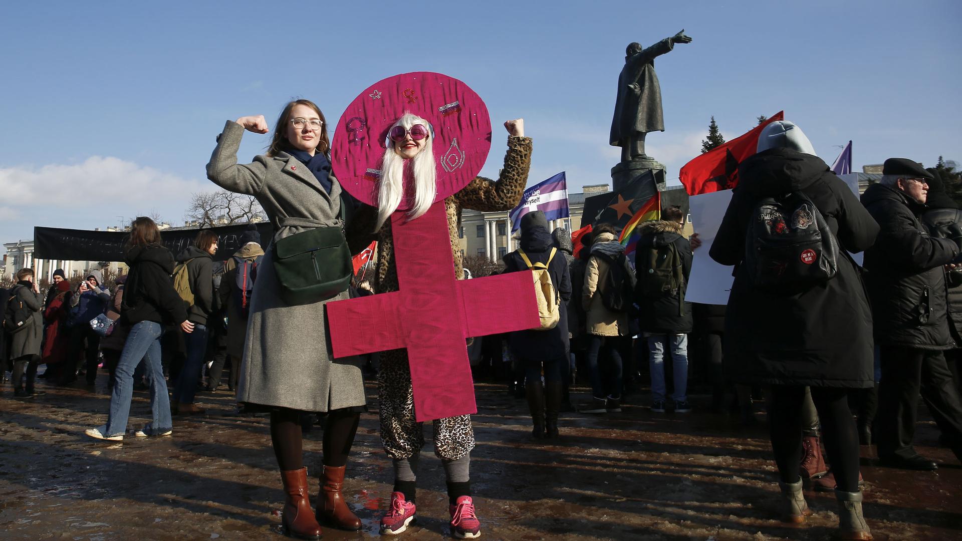 Two women hold their arms in a bicep flex pose. One is wearing a pink cardboard costume in the shape of the women's gender sign. 