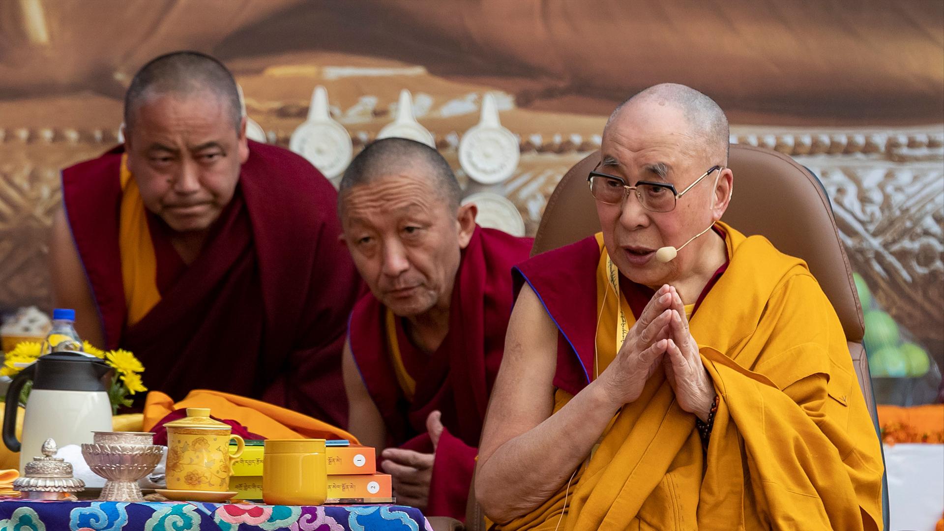 The Dalai Lama speaks while wearing a yellow robe. He is surrounded by two man on red robes in a monastery.