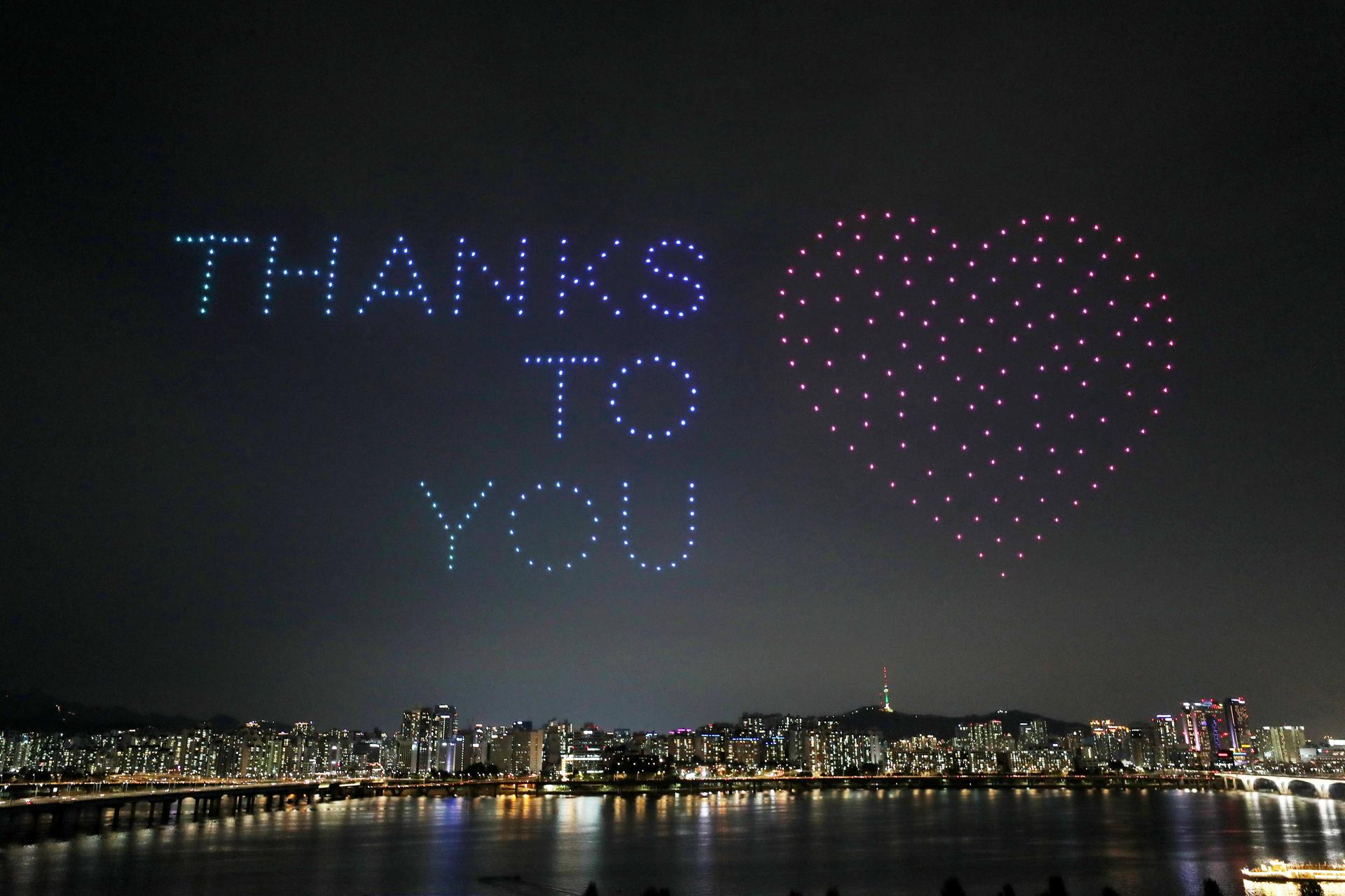 The words Thanks to you are drawn in the sky over a city by lights on drones.