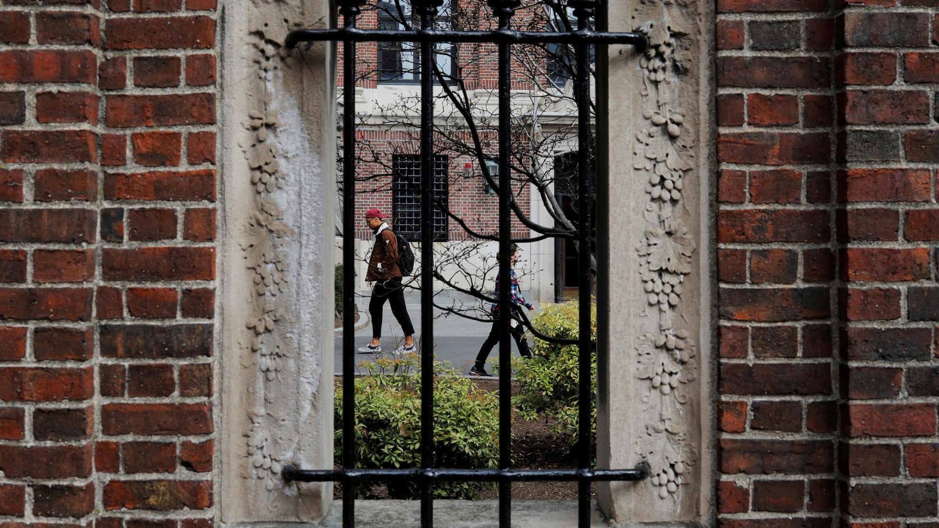 Several people are shown through a stone window with metal bars on it walking.