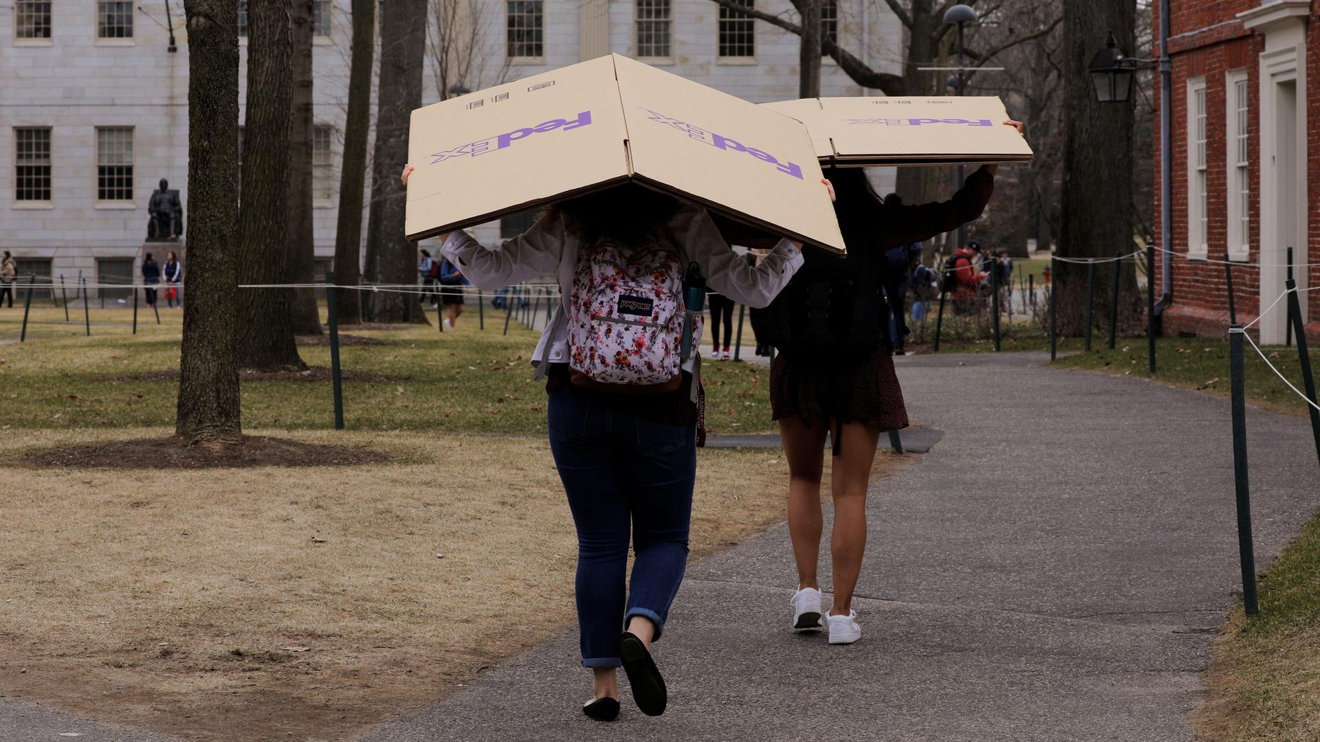 Two women are shown carrying cardboard FedEx boxes over their heads as they walk through Harvard University's campus.