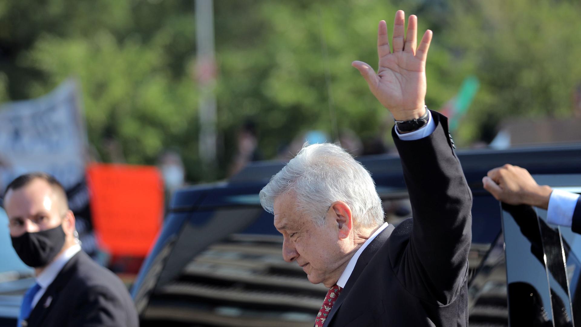 Mexico's President Andrés Manuel López Obrador is shown from the side wearing a dark suit and waving his left hand in the air.