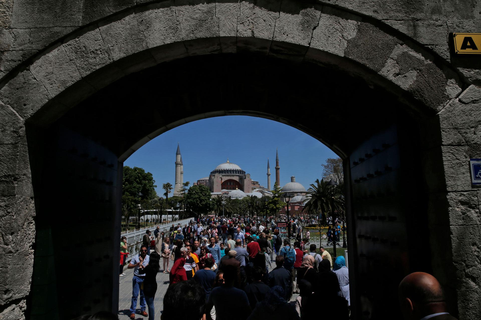 A large crowd of people are shown through an open under a stone passageway with the iconic Hagia Sophia in the distance.