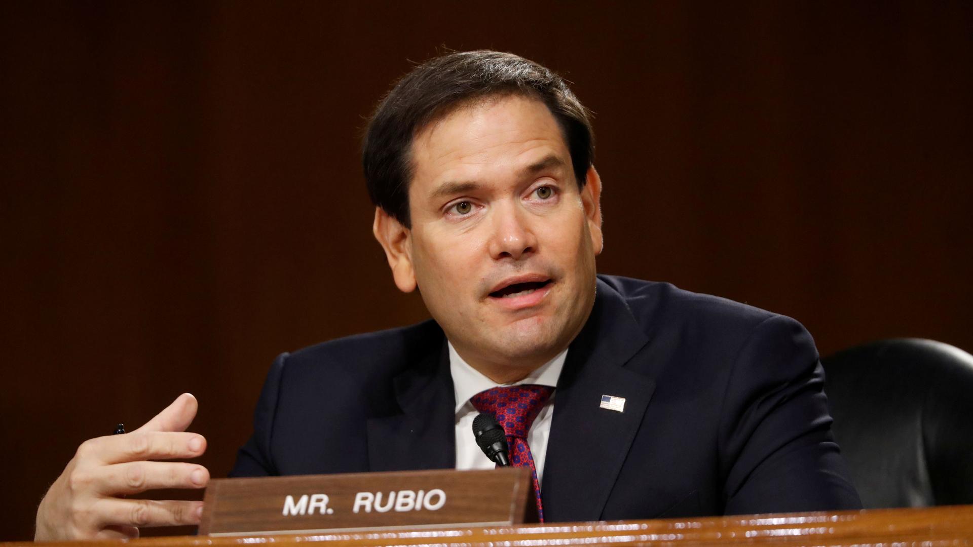 US Sen. Marco Rubio is shown wearing a dark suit and speaking with a small placard in front of him with his name on it.