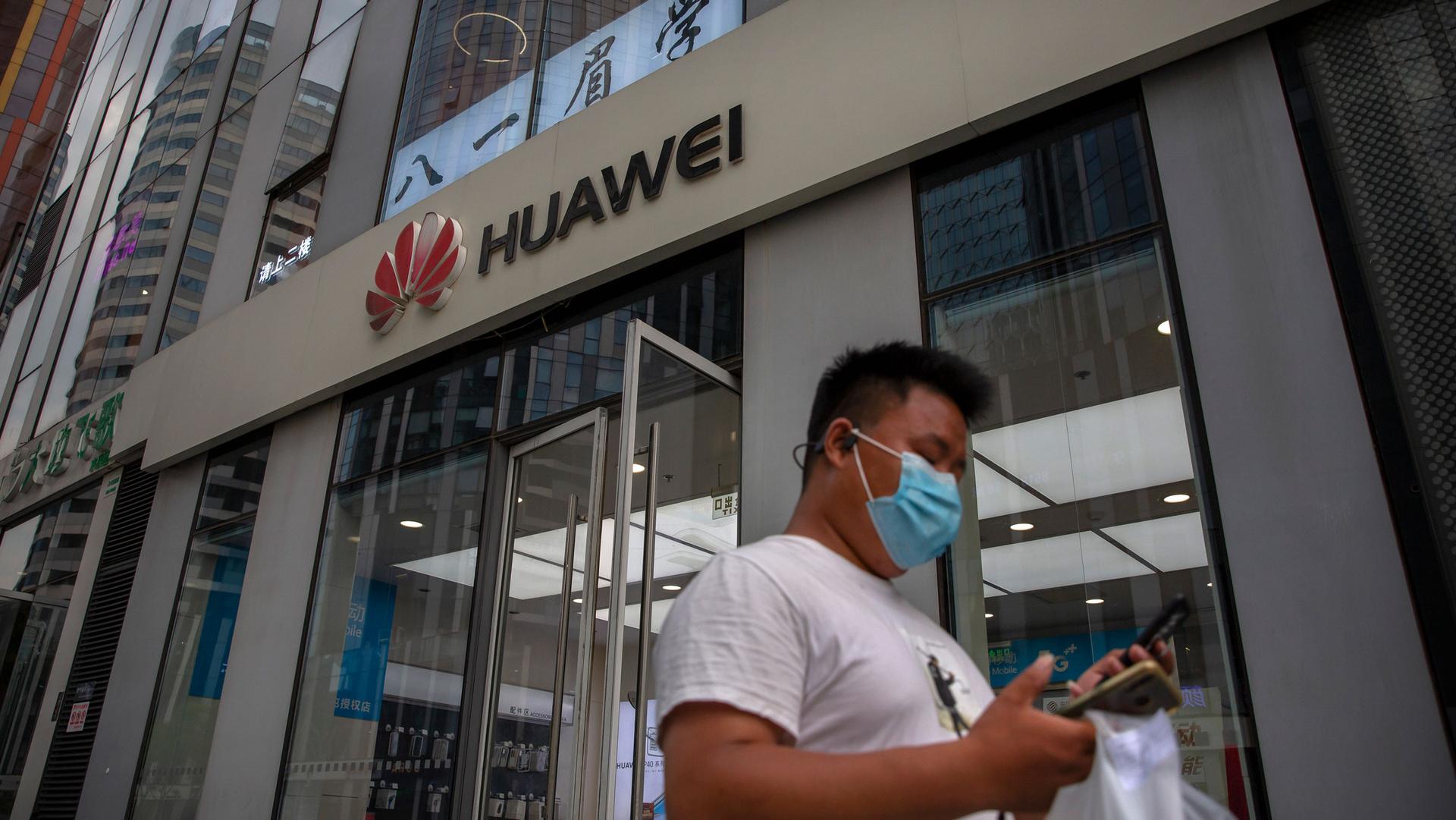 A man is shown looking down at this phone and wearing a face mask with a Huawei store in the background.