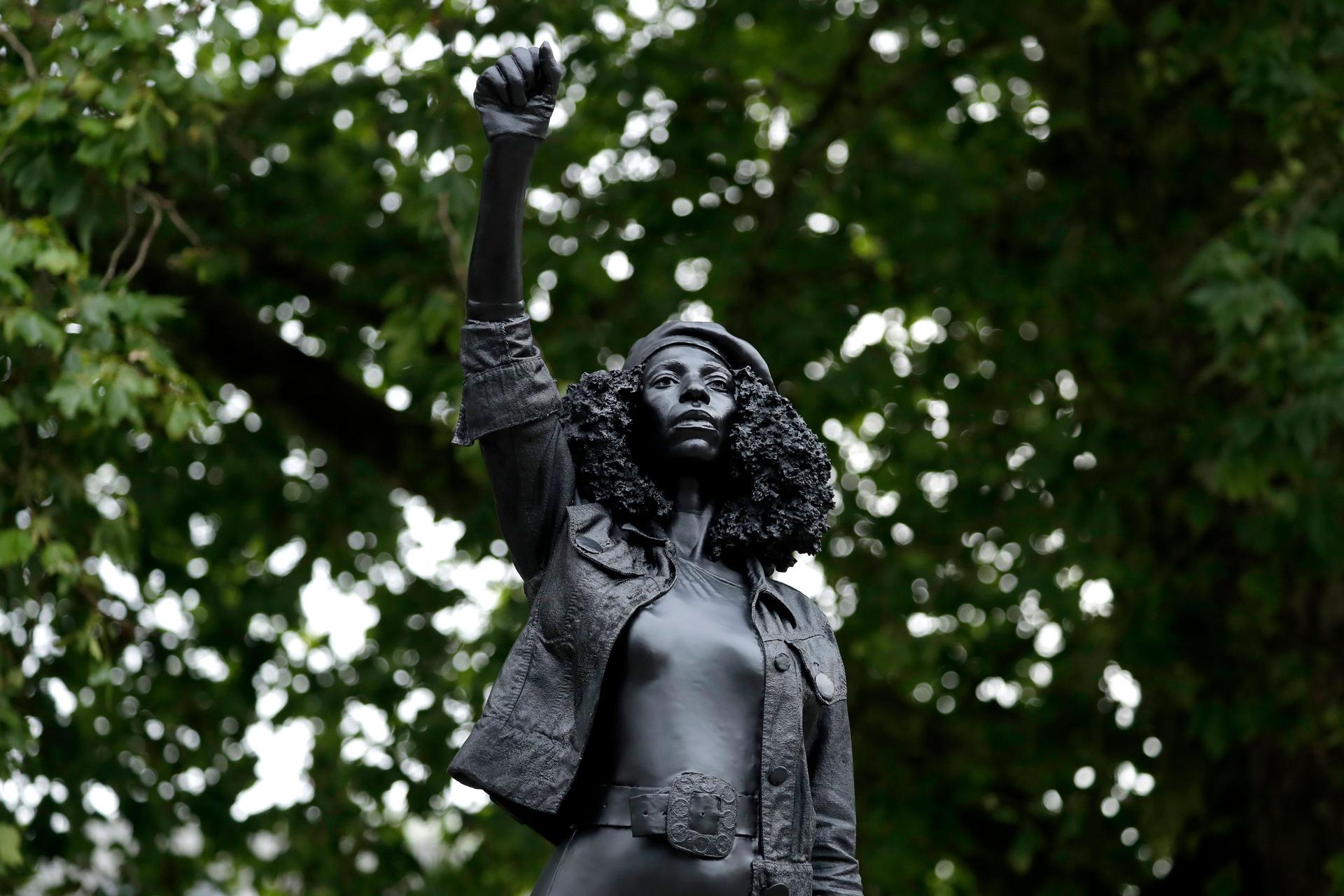 A close-up of a dark statue showing a woman with curly hair, wearing a hat and holding her right fist in the air.
