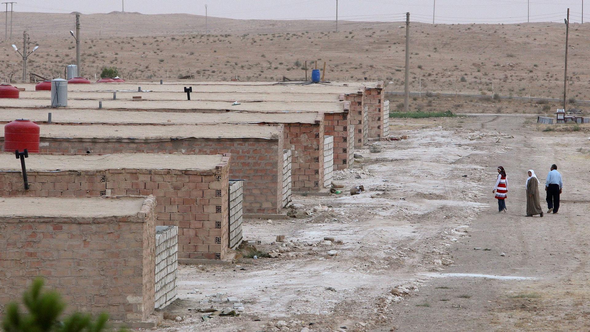 A row of brick shelters are shown amidst a sandy desert location with three people walking in the distance.
