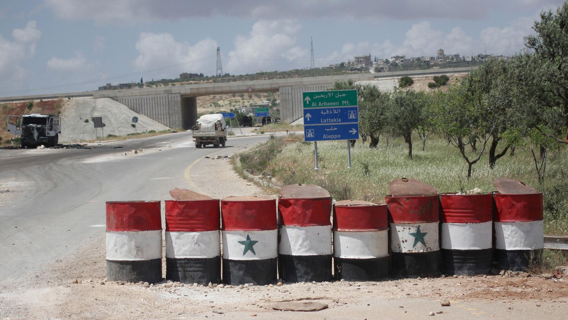 A road is shown with a row of large barrels with the colors of the Syrian flag painted on them.