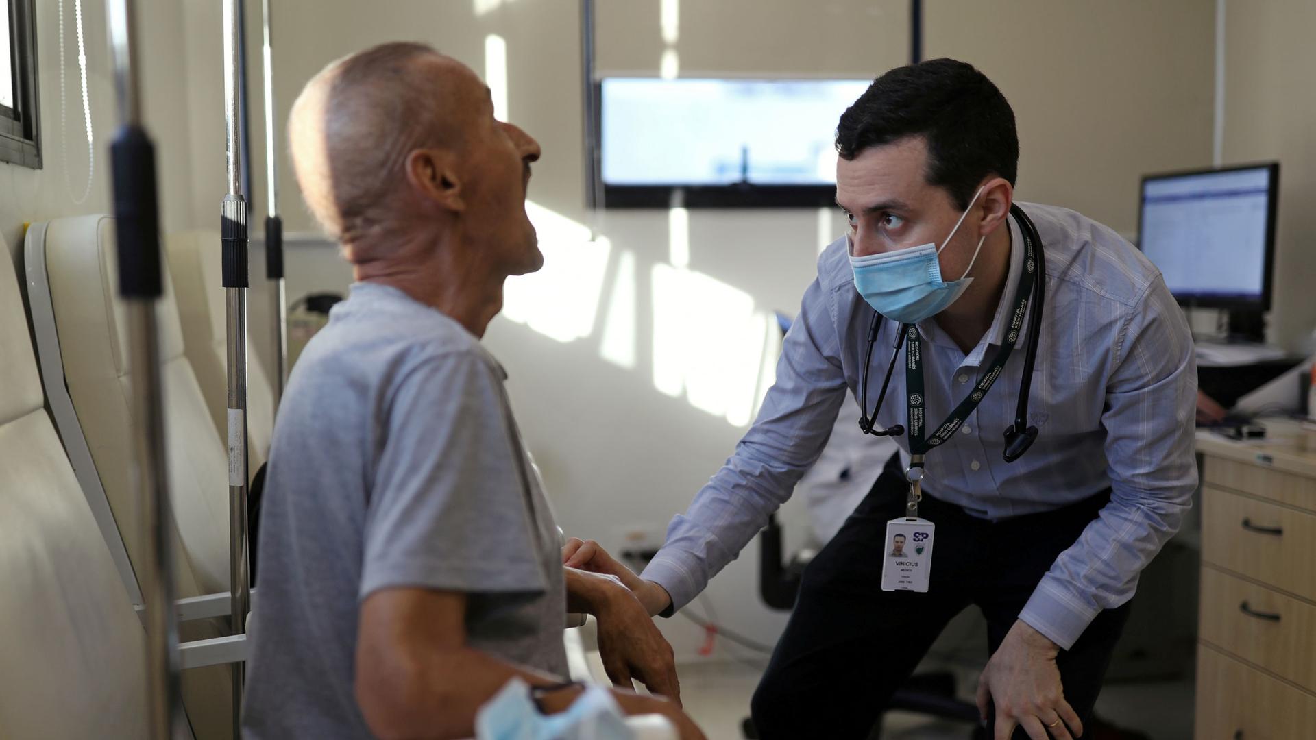 An older man is shown seated and with his mouth open wide with a doctor wearing a face mask looking into his mouth.