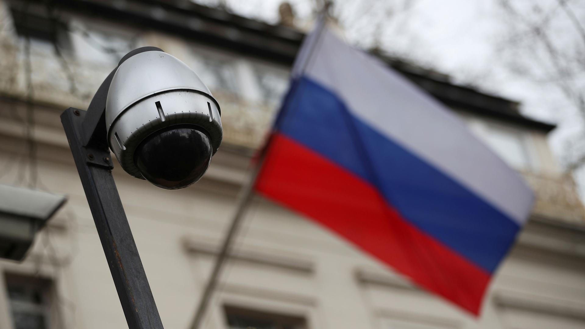 The dark lens of a security camera is shown with a Russian Federation flag in soft focus in the background.