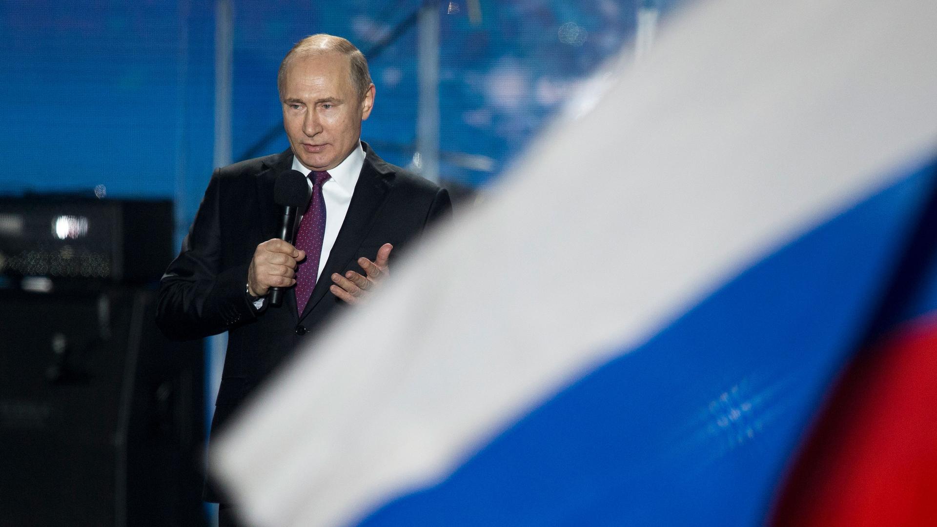 Russian President Vladimir Putin is shown speaking into a microphone and wearing a dark suit.