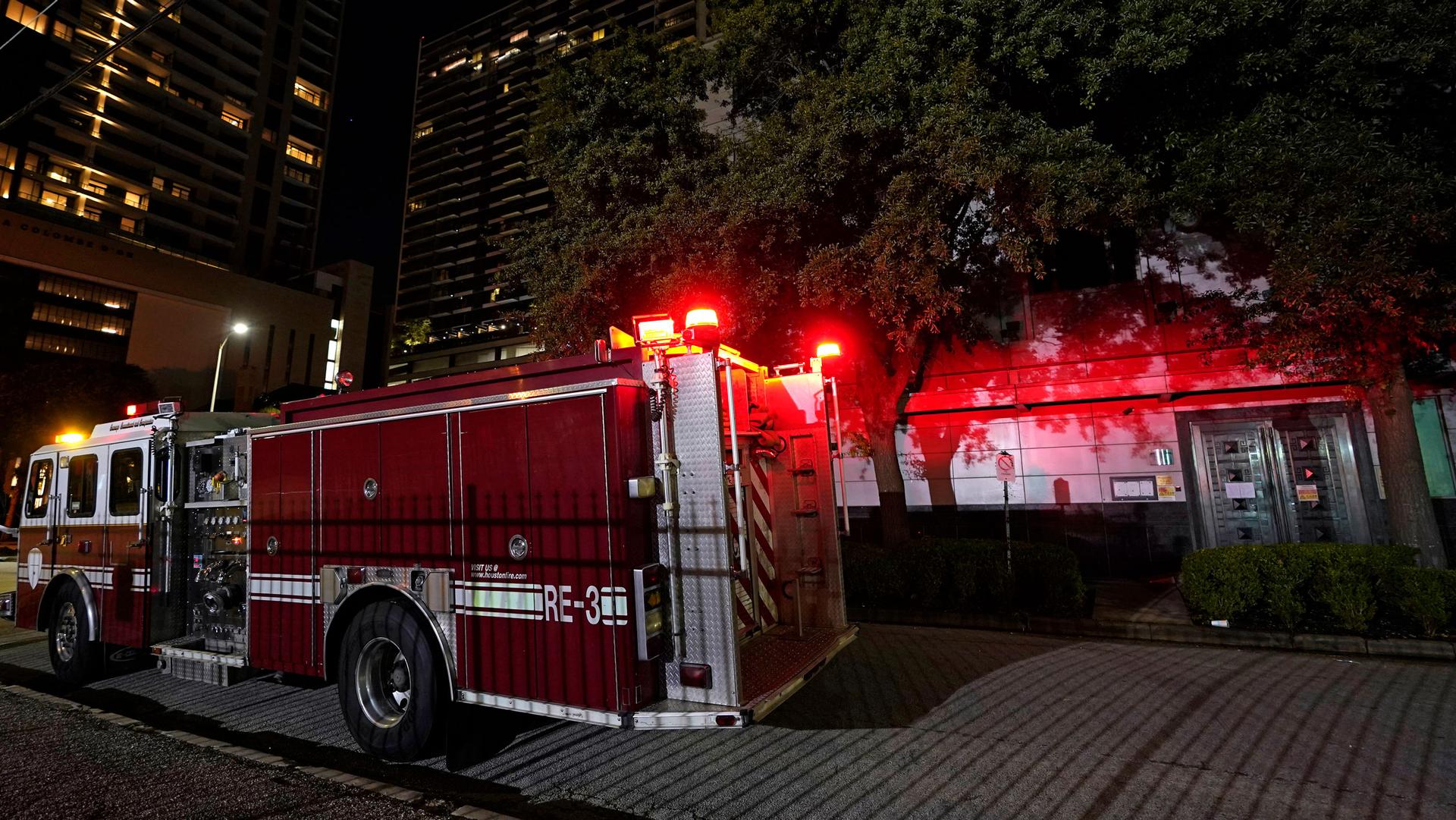 The rear of a red fire truck is shown with lights on outside of a building with trees in the background.