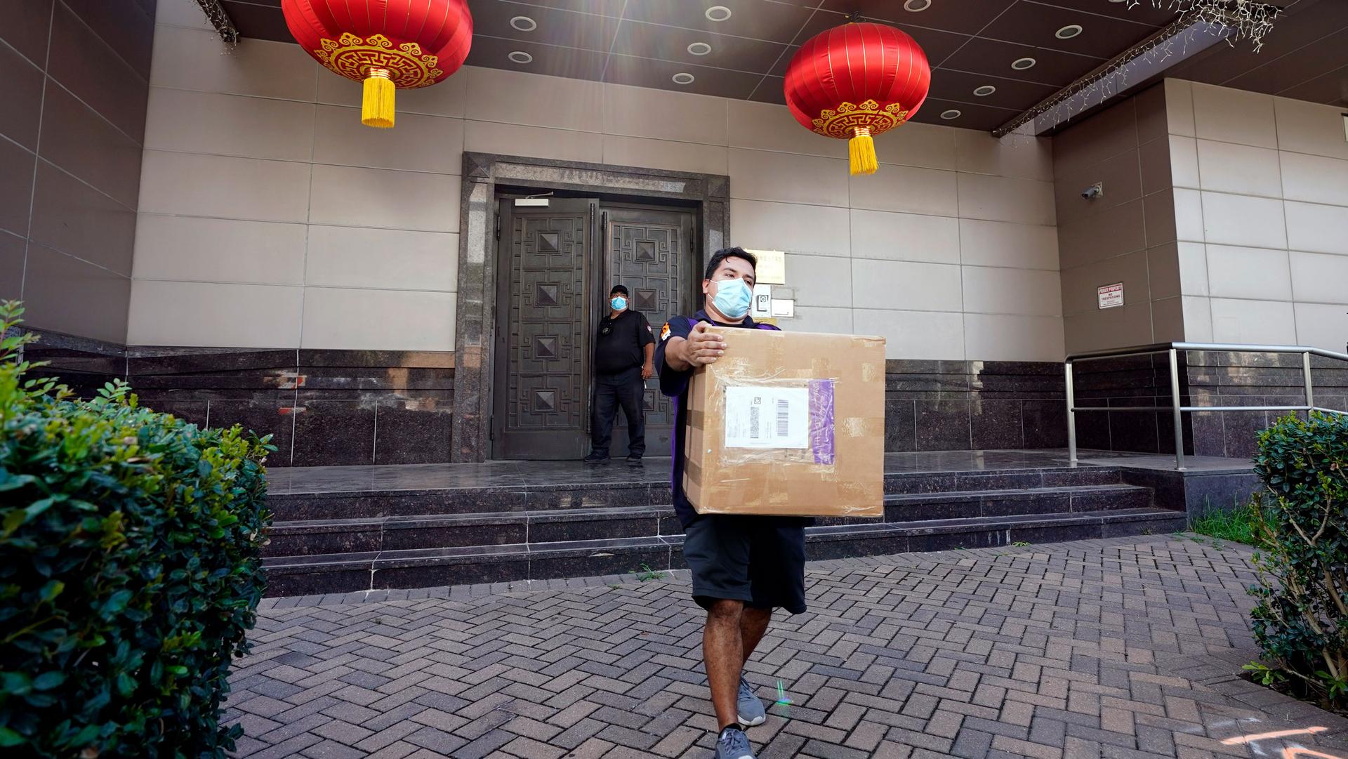 A man is shown wearing a face mask and carrying a large brown cardboard box with the doors of the Chinese Consulate in Houston in the background.