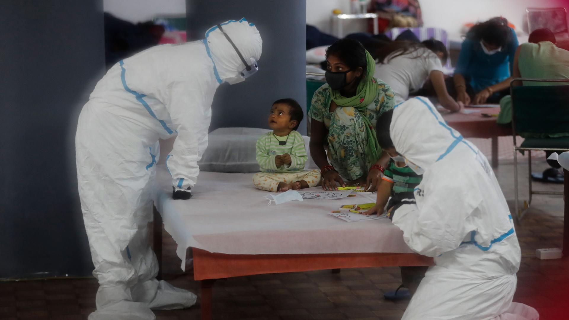 A young child is shown sitting on a bed with health care workers nearby dressed in full protective suits.