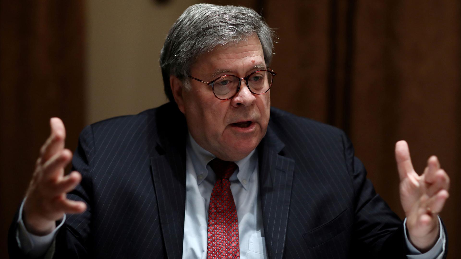 US Attorney General William Barr is shown wearing a dark suit and red tie with his hands outstretched.