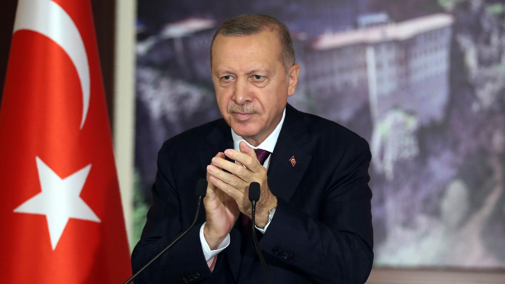 Turkey's President Recep Tayyip Erdoğan is shown wearing a dark suit and standing behind a microphone with a Turkish flag nearby.