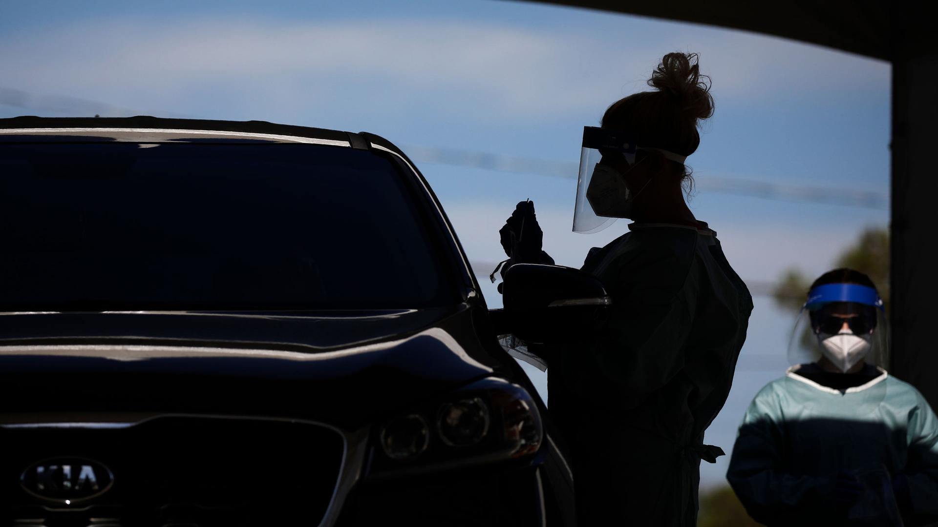 A healthcare worker is shown in shadow standing next to a car and wearing a protective face shield.