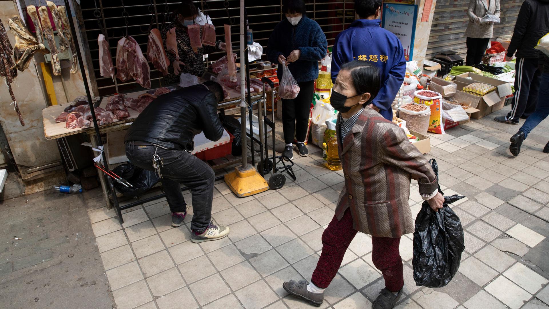 Several people are shown outside of a partially closed market stall with several pieces of meat hanging in the background.