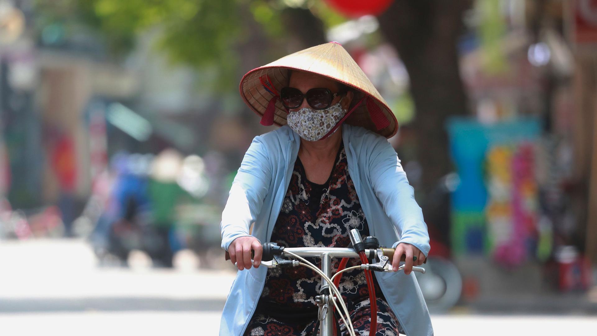 A woman is shown wearing a blue jacket, sunglasses, face mask and a straw hat while writing a bicycle.