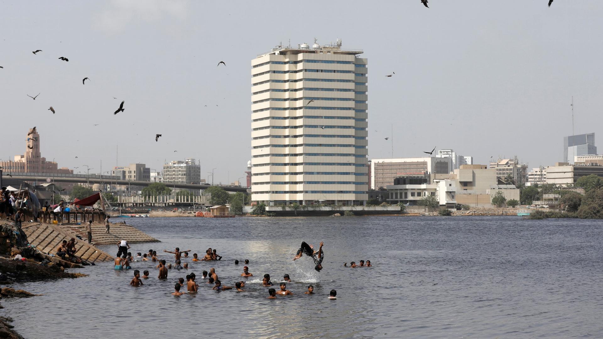 Adults and children swim in water near a large, white building in the background and birds flying overhead