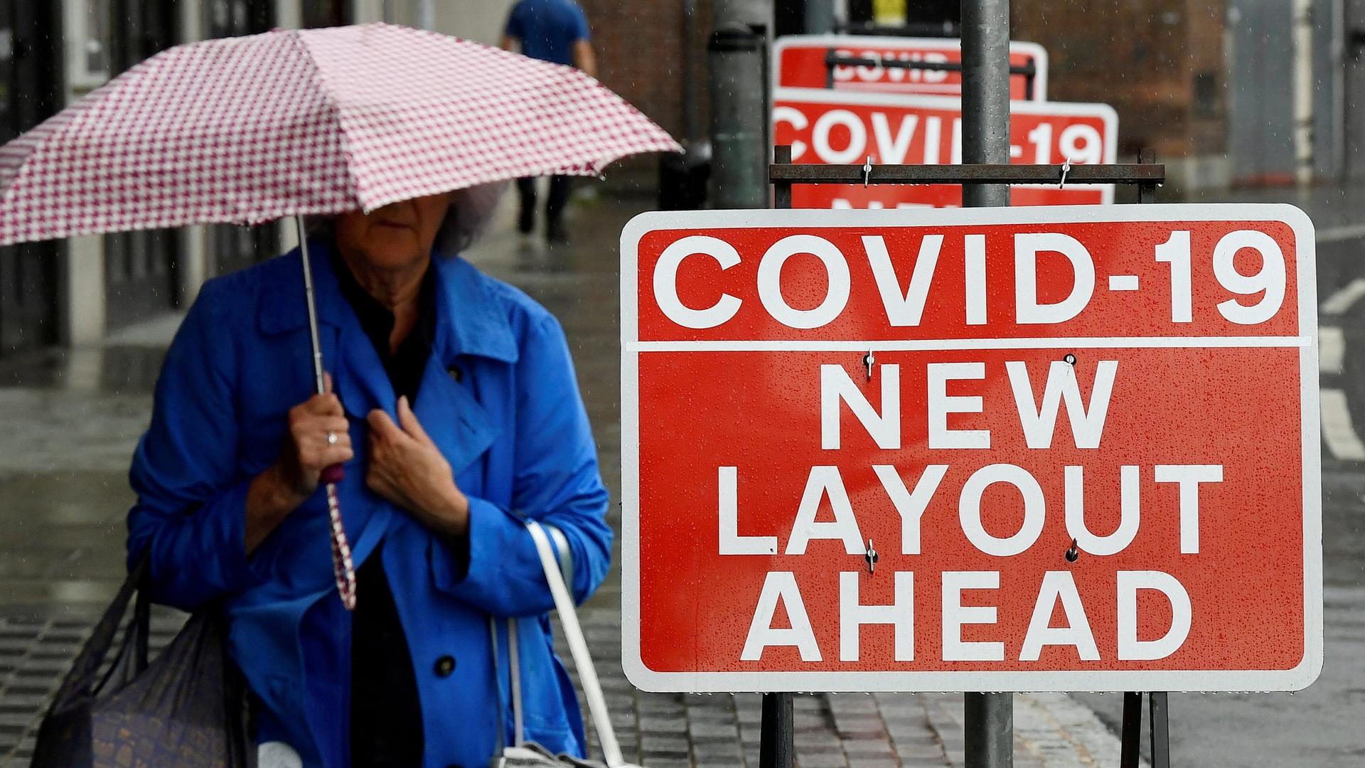A woman holds shopping bags and an umbrella next to bright red sign with white text about COVID-19