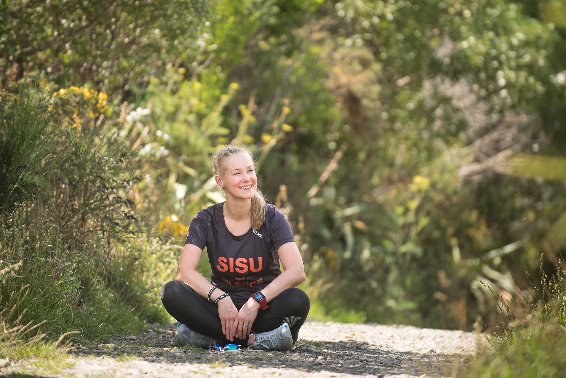 A white woman sits near lots of greenery smiling and wearing black clothes with the word sisu in red letters on her shirt.