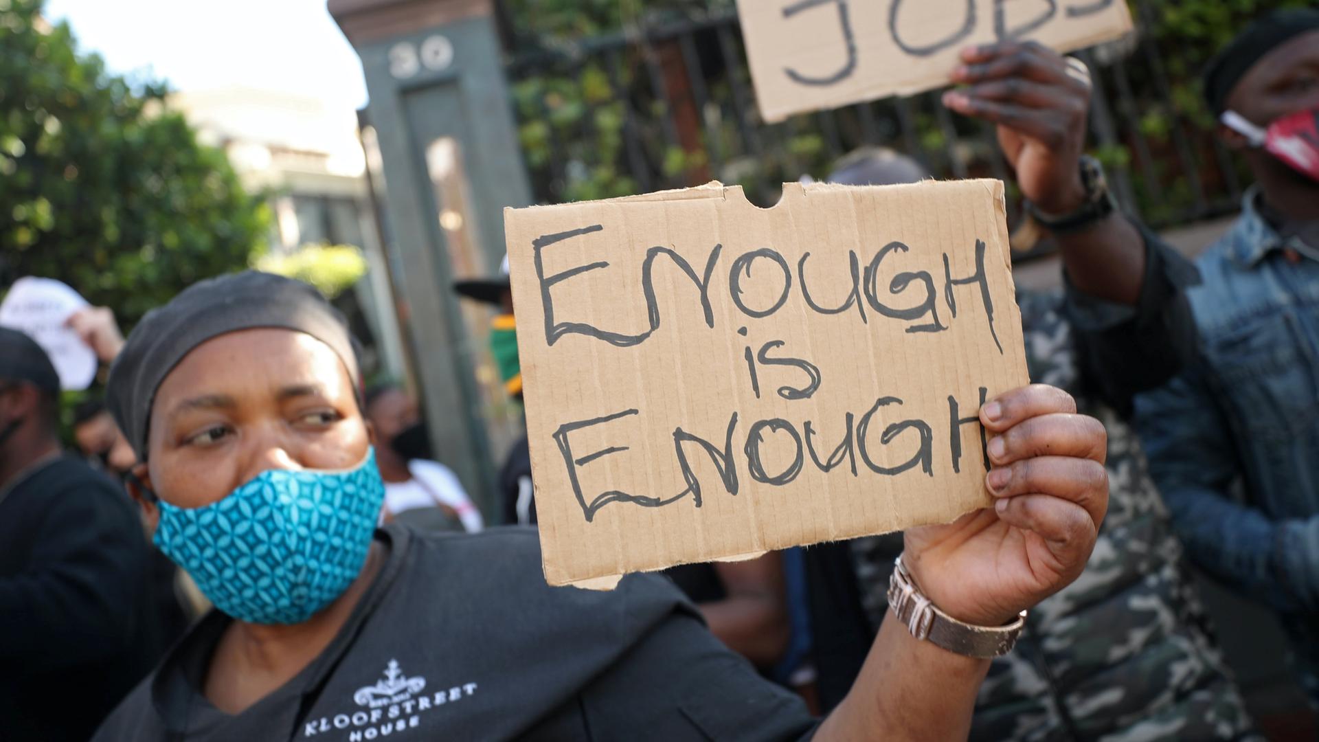 A Black African woman wears a blue face mask and holds a brown sign that reads "enough is enough."