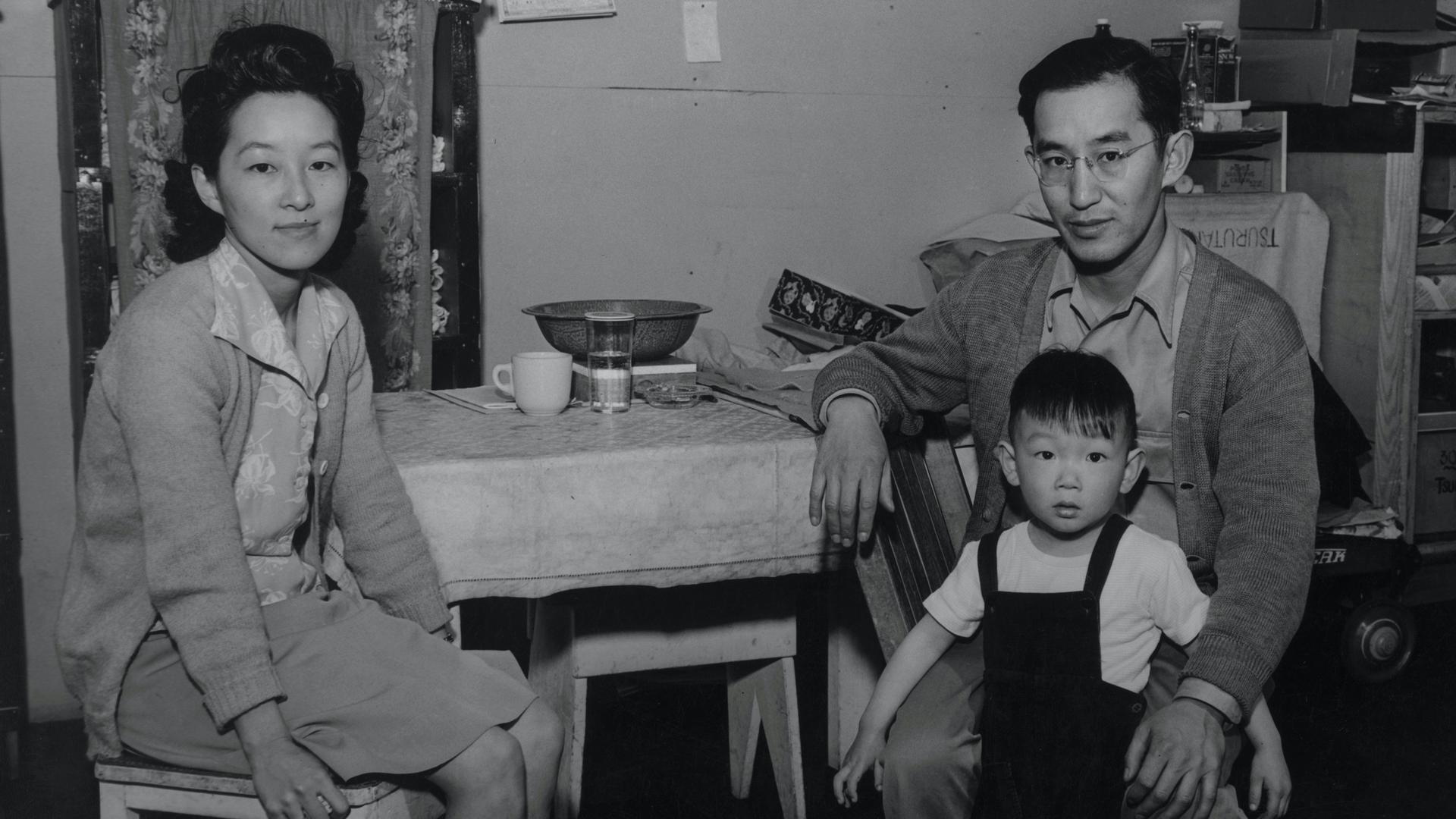 A black and white photo of a Japanese family (woman, man, child) sitting in a kitchen setting.