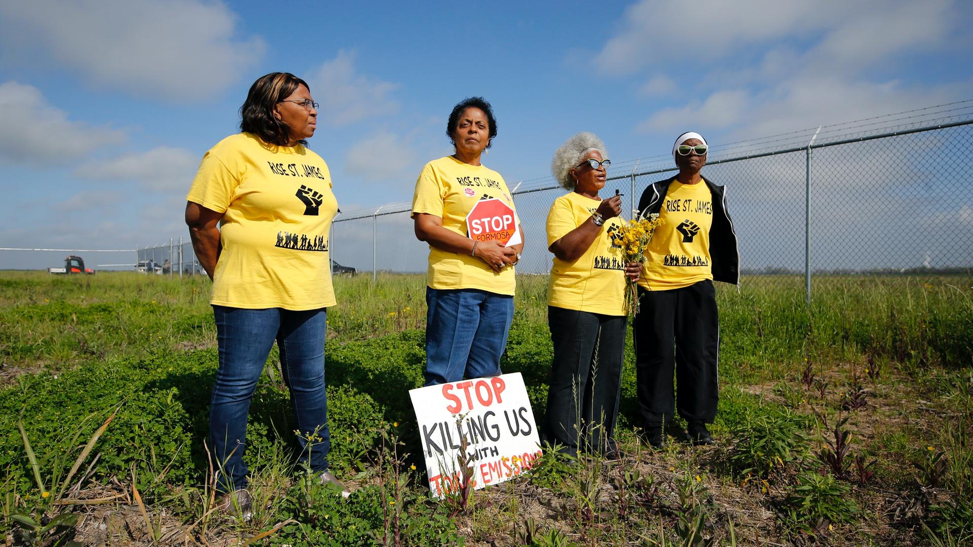 Four Black American women activists wearing yellow shirts and holding protest signs stand in a field near a fence. 