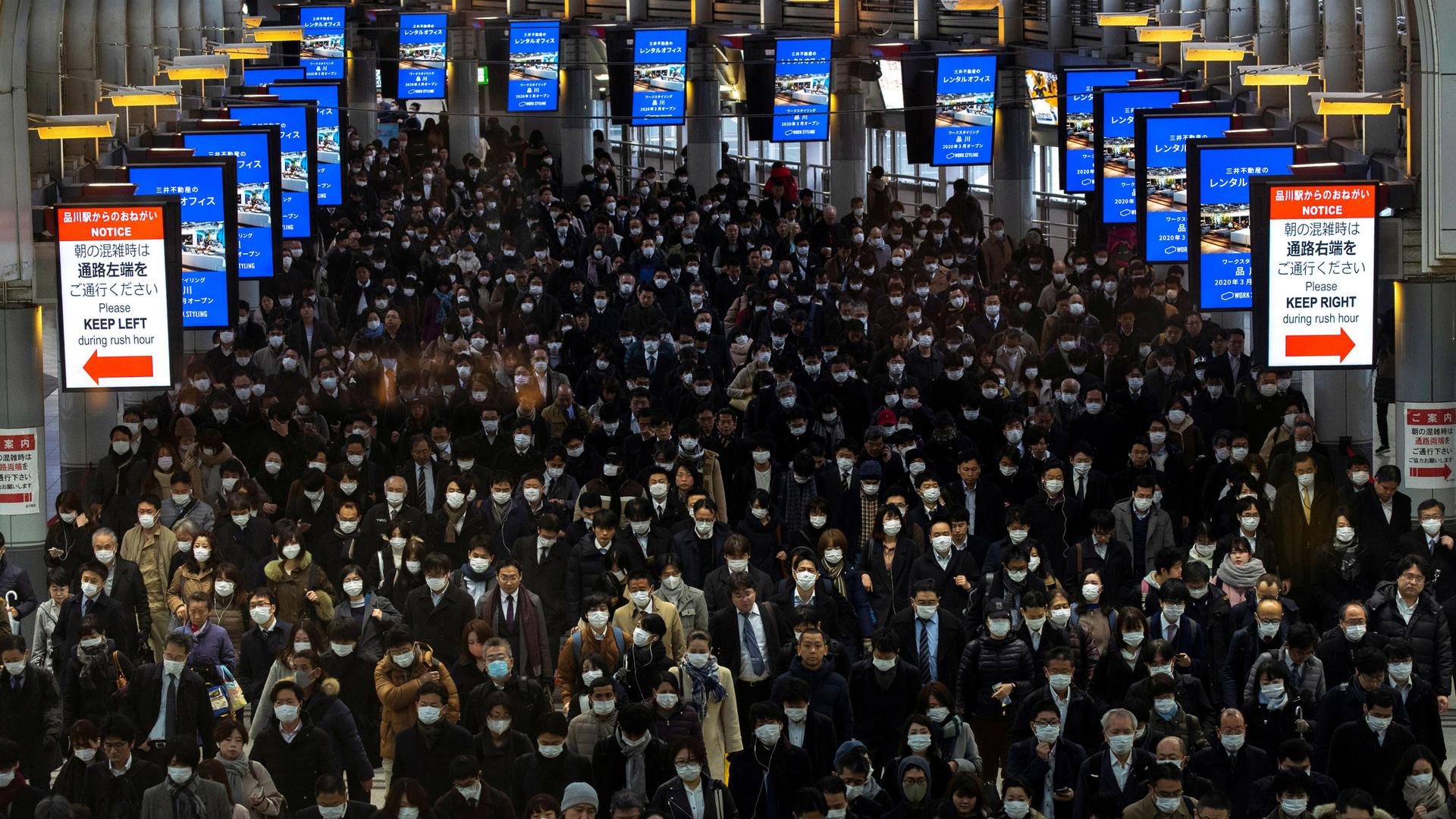 A large open area of a train staton is shown crowded with people most of which are wearing protective face masks.