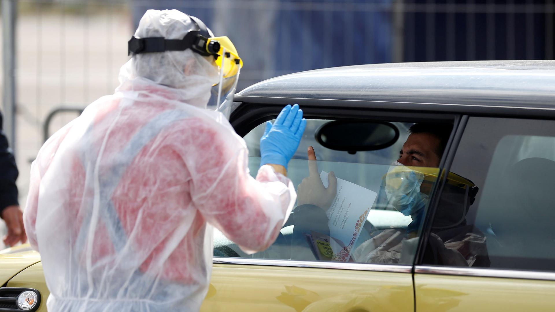A health worker wearing a full protective gown and face guard is shown standing with his hand in the air at a drive-through coronavirus disease testing center.