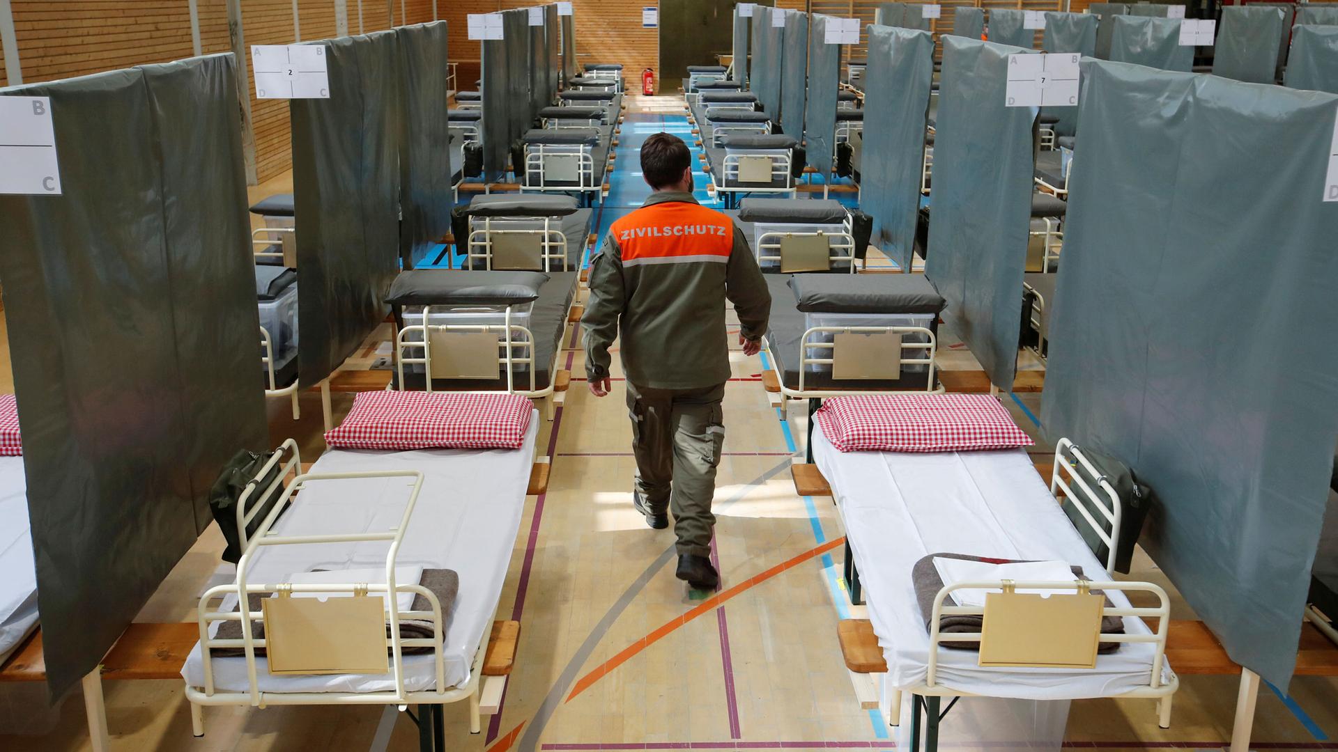 A man wearing a work uniform with an orange top walks down an aisle with several beds lining both sides.