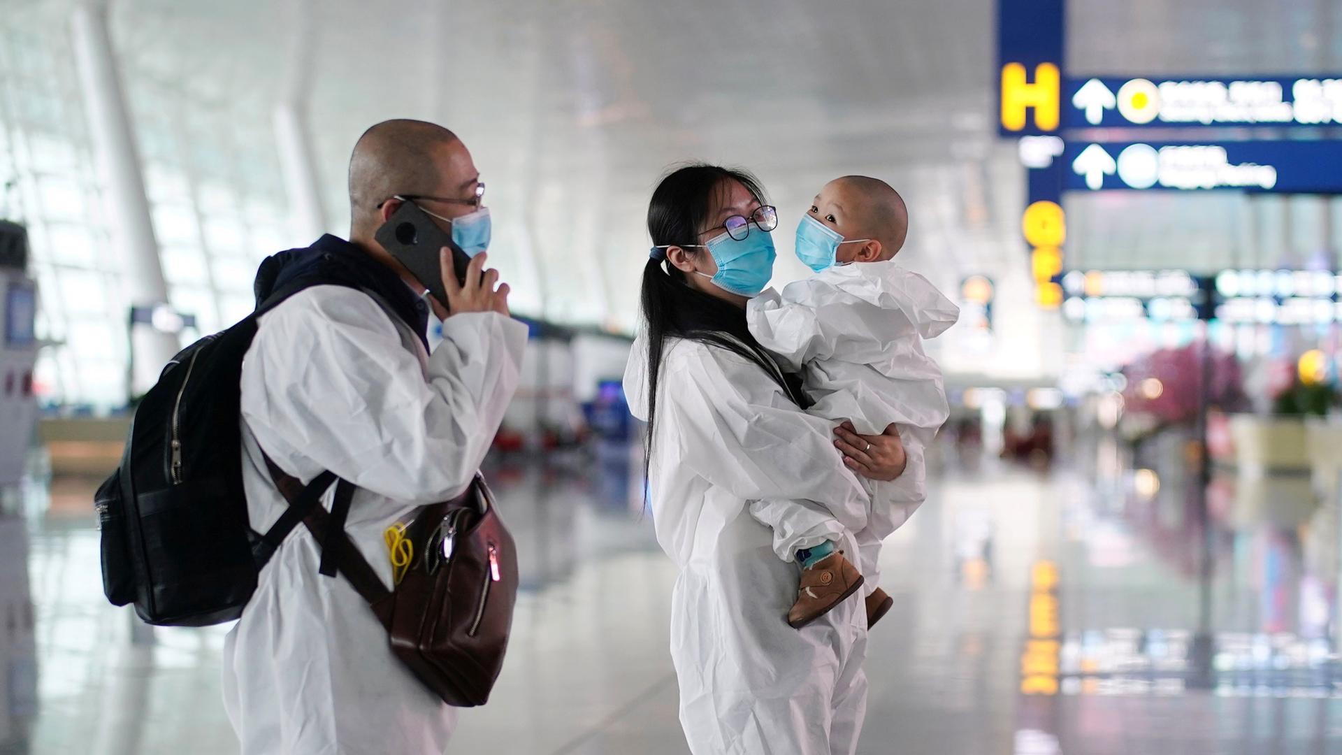 Travelers in white protective suits and face masks are shown, with one talking on a mobile phone and the other holding a small child.